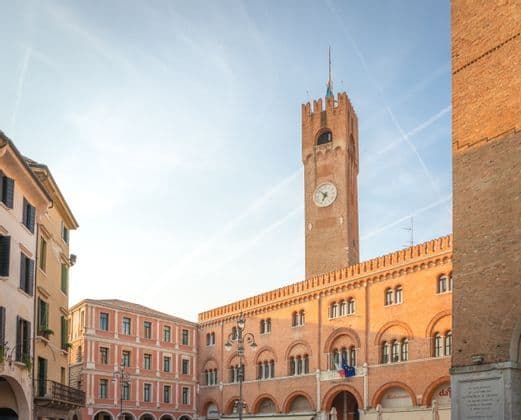 Ein sonnenbeschienener, roter Backsteinglockenturm und ein angrenzendes Bogengebäude stehen auf einem historischen Marktplatz unter einem hellblauen Himmel.
