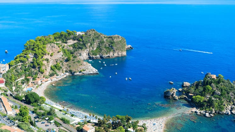 Vista aerea di una baia costiera con una spiaggia sabbiosa collegata a una piccola isola rocciosa e alberata, in acque cristalline.
