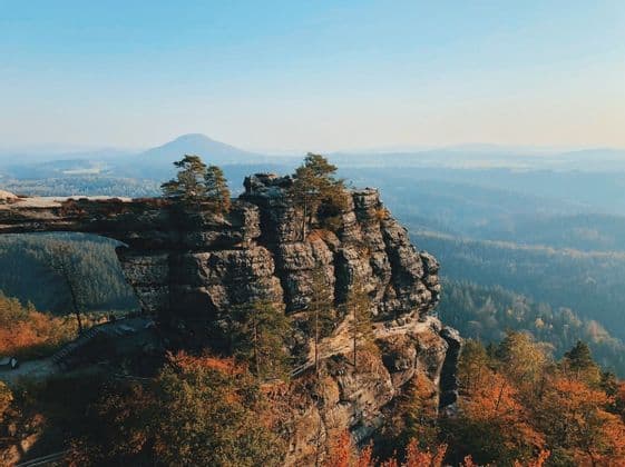 Ein natürlicher Steinbogen auf einer felsigen Klippe überblickt ein weites Tal mit Wäldern in Herbstfarben und fernen, dunstigen Bergen.