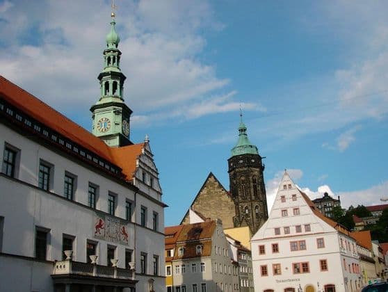 Über historischen Gebäuden mit roten Ziegel- und Giebeldächern auf einem europäischen Stadtplatz erhebt sich ein Uhrturm mit grünem Turmhelm.