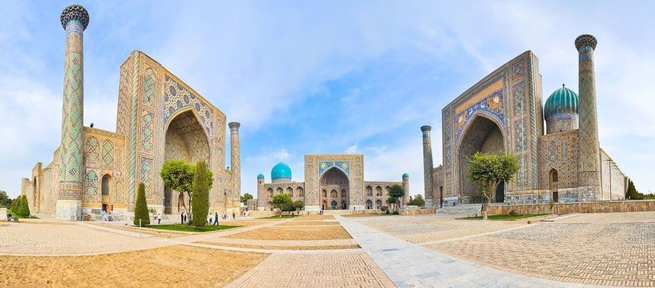 Vue panoramique sur une grande place historique, flanquée de trois bâtiments ornés, avec des mosaïques complexes, de hauts minarets et des dômes bleus.