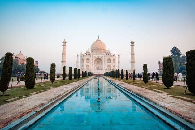 Il Taj Mahal si riflette in una lunga piscina fiancheggiata da giardini con cipressi, con visitatori che passeggiano sui sentieri.