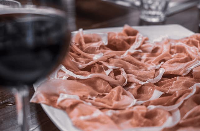 Un primo piano di un piatto bianco colmo di prosciutto crudo tagliato finemente, con un bicchiere di vino rosso sfocato in primo piano.