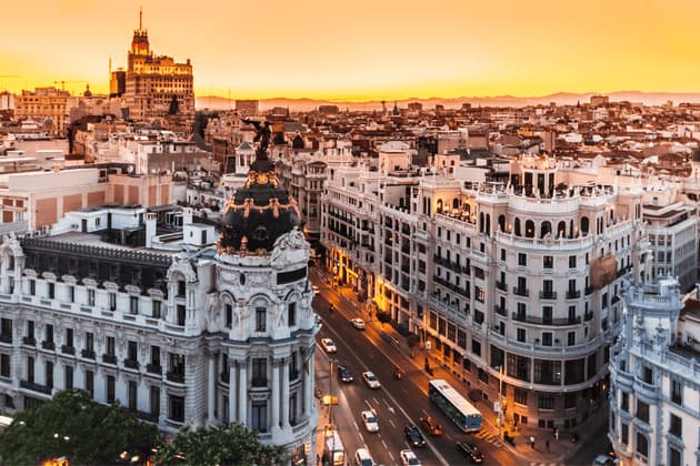 Une vue aérienne d'une ville animée avec une architecture ornée et des rues remplies de trafic, le tout sous un coucher de soleil orange éclatant.