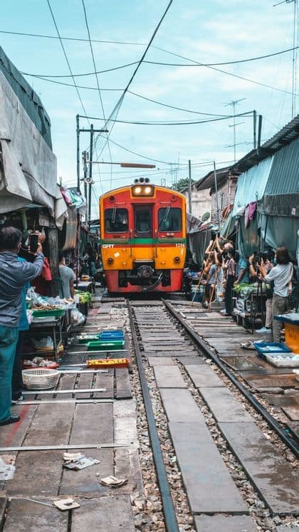 Un treno arancione e rosso si avvicina sui binari che attraversano un mercato affollato mentre le persone si spostano per scattare foto.