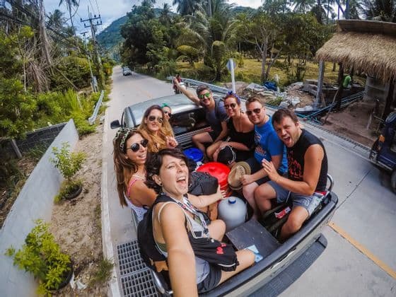 Un voyage en groupe WeRoad prenant un selfie en souriant à l'arrière d'un pick-up sur une route bordée de palmiers.