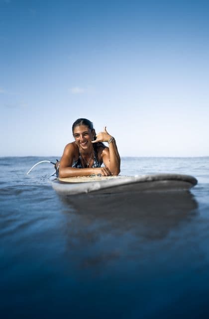 Une femme allongée sur sa planche de surf dans l'océan, souriante et faisant un signe shaka vers la caméra.