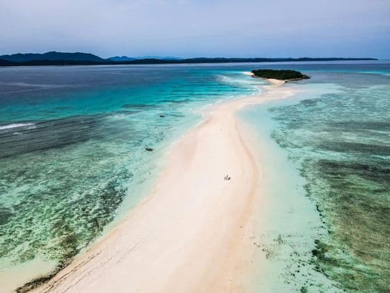 Una veduta aerea di un gruppo WeRoad in cammino su una lunga lingua di sabbia bianca che si protende in un mare turchese verso una piccola isola.