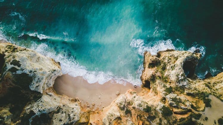 Vue aérienne de vagues turquoise de l'océan s'écrasant sur une plage de sable isolée, entourée de hautes falaises rocheuses.