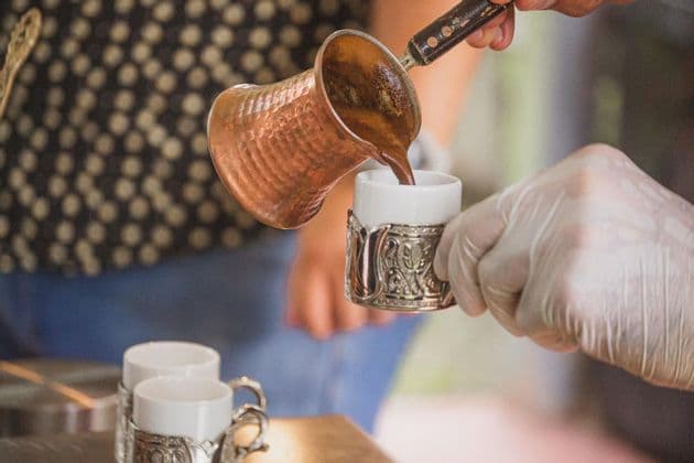 Una persona vierte café caliente de un cezve de cobre tradicional en una pequeña taza blanca sostenida por una mano enguantada.