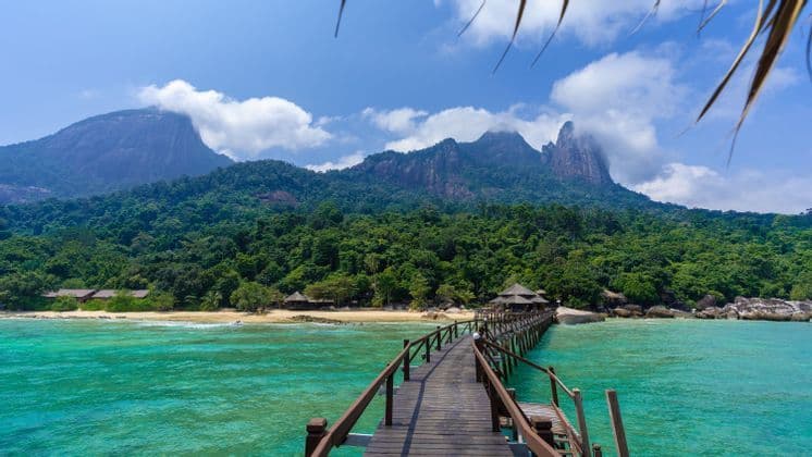Un lungo molo di legno si estende su acque turchesi verso una spiaggia sabbiosa, con dietro montagne verdi e lussureggianti sotto un cielo azzurro con nuvole soffici.