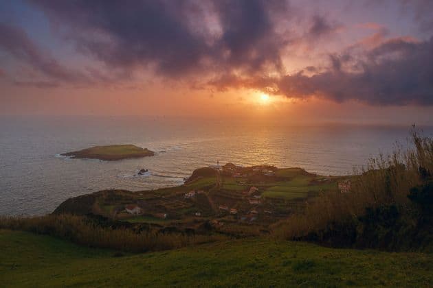 Una vista dall'alto di un villaggio costiero e di una piccola isola nell'oceano, durante un tramonto spettacolare con nuvole arancioni e viola.