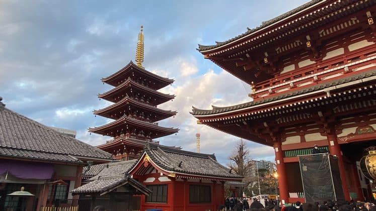 Una pagoda rossa a cinque piani e un grande portale del tempio si ergono maestosi contro un cielo nuvoloso, con una folla di persone radunate sotto.