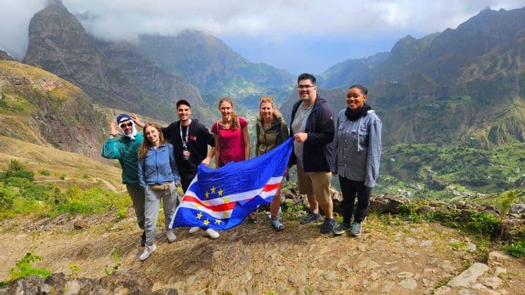 Sieben Personen einer WeRoad Gruppenreise posieren mit einer Kapverden-Flagge auf einem Bergpfad mit Blick auf ein grünes Tal.