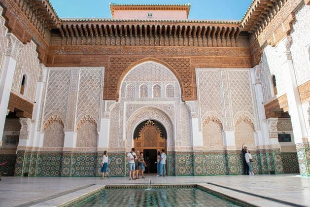 A WeRoad group trip standing in a sunny, ornate courtyard with a central pool, intricate tilework, and carved plaster walls.