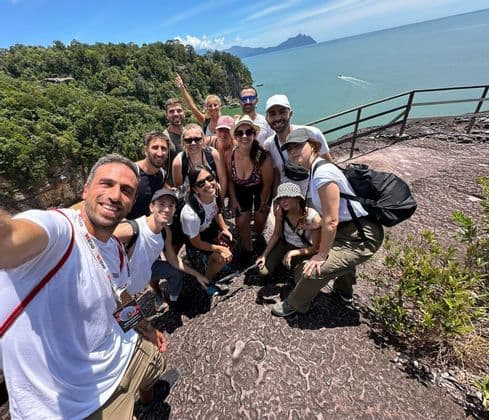 Un gruppo WeRoad in viaggio posa per un selfie di gruppo su una scogliera rocciosa che domina una costa boscosa e il mare.
