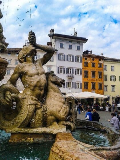 Una fontana in pietra con scultura di tritone che spruzza acqua in una piazza europea con edifici colorati.