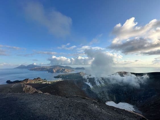 Il vapore si innalza da un cratere vulcanico che domina una città costiera e isole lontane nel mare, sotto un cielo parzialmente nuvoloso.