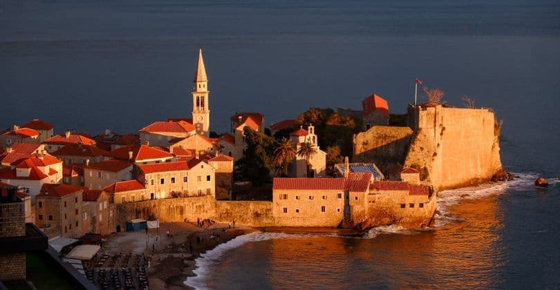 Eine befestigte Küstenstadt mit roten Ziegeldächern und einem markanten Glockenturm wird von goldenem Sonnenuntergangslicht neben dem dunklen Meer beleuchtet.