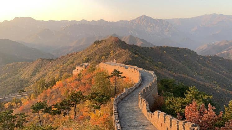 Un lungo muro di pietra si snoda attraverso dolci colline coperte da un fogliame autunnale colorato sotto un cielo velato al tramonto.