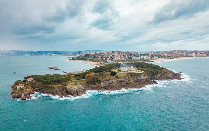 Vista aérea de un palacio en una península arbolada, rodeado por el mar, con una ciudad costera al fondo bajo un cielo nublado.