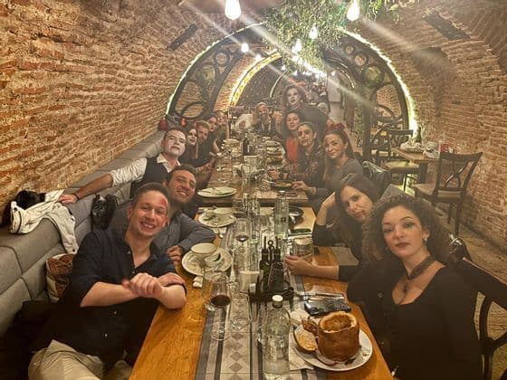 Un viaggio di gruppo WeRoad in costume di Halloween che sorride per una foto attorno a un lungo tavolo da cena in un ristorante in cantina con volte in mattoni.