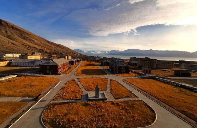 Una vista aérea de un asentamiento remoto con edificios uniformes, situado entre una montaña árida y un fiordo con glaciares distantes.