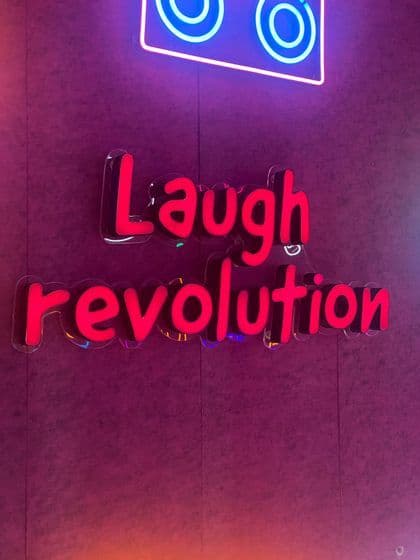 Un letrero iluminado con las palabras 'Revolución de la risa' en letras rojas, montado sobre una pared morada texturizada bajo una luz de neón azul.