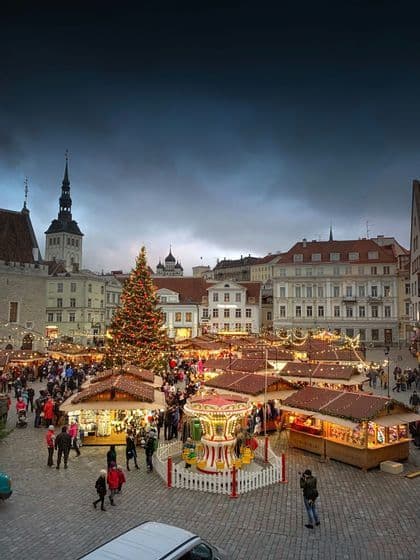 Un vivace mercatino di Natale anima una piazza lastricata con un grande albero decorato, bancarelle e una giostra al crepuscolo.