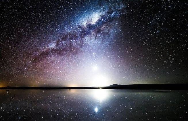 The Milky Way and countless stars in the night sky perfectly reflect onto a calm body of water on the horizon.