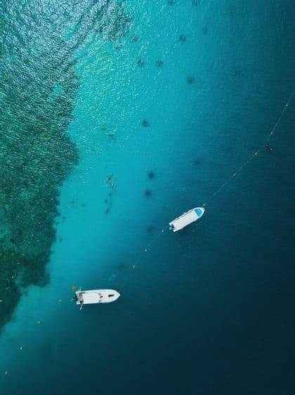 Una vista aerea di due barche bianche ormeggiate in acqua turchese, con diverse persone che fanno snorkeling nelle vicinanze sopra una barriera corallina.