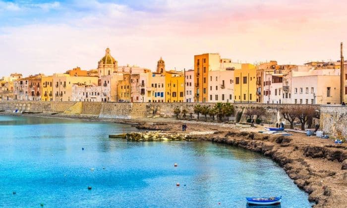 Una cittadina costiera con edifici colorati si affaccia su una diga di pietra lungo acque di un blu brillante, sotto un cielo dalle sfumature pastello.