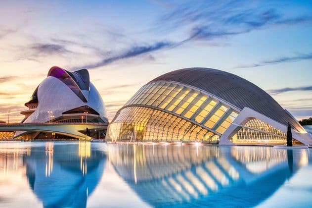 Due moderni edifici illuminati si riflettono in uno specchio d'acqua calmo, sotto un cielo vibrante al tramonto.