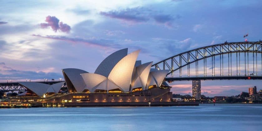 A modern building with white, sail-shaped roofs stands illuminated next to a large arch bridge over the water at dusk.