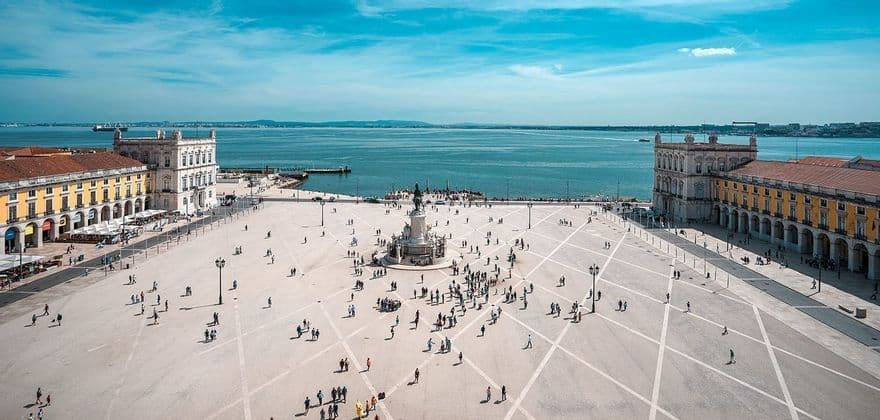 Una vista aérea muestra a personas caminando en una gran plaza pavimentada junto al mar con un monumento central, flanqueada por edificios porticados bajo un cielo azul.