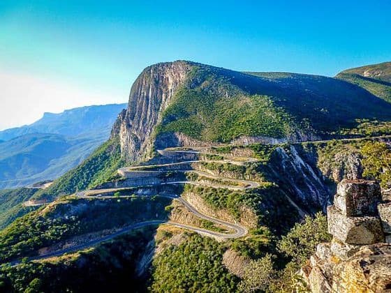 Una strada tortuosa con numerosi tornanti si inerpica su una montagna verde e lussureggiante sotto un cielo azzurro e limpido.