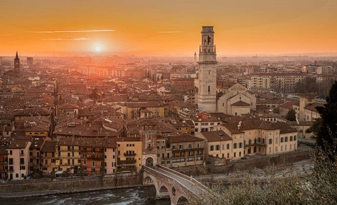 Una veduta aerea di una città storica con un ponte di pietra e un campanile sotto un tramonto arancione splendente.