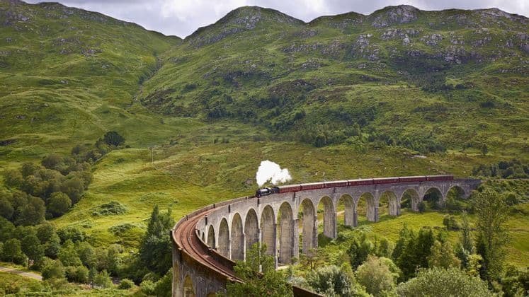 Un treno a vapore con vagoni rossi attraversa un viadotto in pietra curvo in una rigogliosa valle verde circondata da montagne.