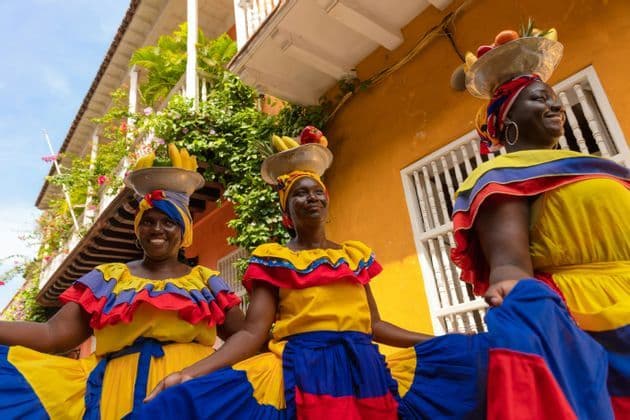 Tre donne sorridenti in abiti tradizionali colorati bilanciano ciotole di frutta sulle loro teste su una strada assolata.