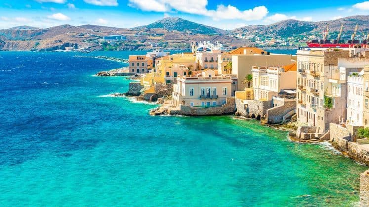 Un borgo marinaro con case colorate si adagia su una costa rocciosa bagnata da un mare turchese, con colline aride sullo sfondo.