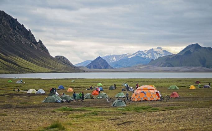 Un voyage de groupe WeRoad avec des tentes colorées et des vélos, campant au bord d'un lac au pied de montagnes escarpées et enneigées.