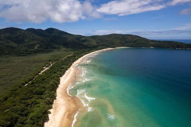 An aerial view of a long, sandy beach where turquoise water meets a dense green forest covering hills under a partly cloudy sky.