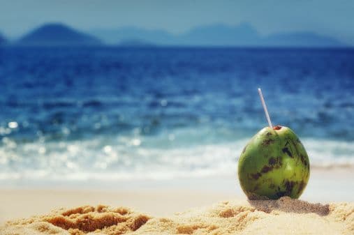 A green coconut with a drinking straw sits in the sand on a beach with the blue ocean in the background.