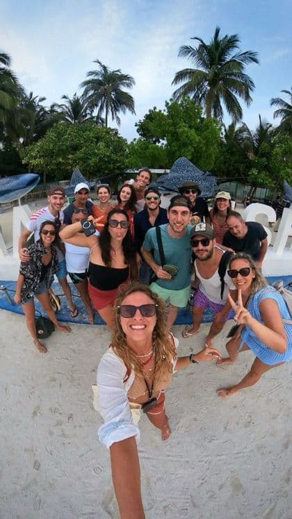 Un gruppo WeRoad si fa un selfie grandangolare su una spiaggia sabbiosa circondata da palme.
