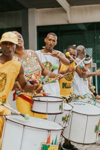 Un gruppo di musicisti in abiti coordinati gialli e bianchi suona energicamente grandi tamburi bianchi in fila.