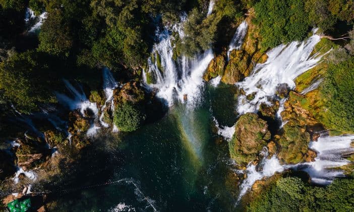 Vista aerea di cascate che si gettano su rocce e vegetazione in una profonda pozza turchese, con un arcobaleno visibile nella nebbia.