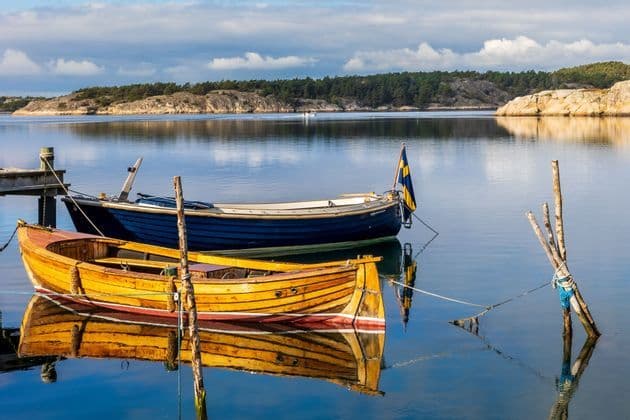 Two wooden boats, one yellow and one blue with a Swedish flag, are moored in calm water with their reflections on the surface.