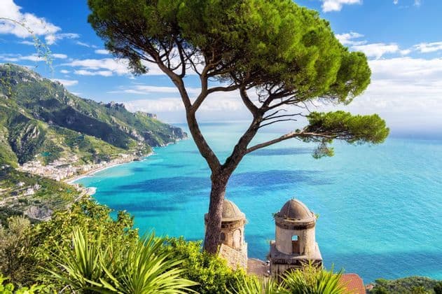 Un pino piñonero en un acantilado enmarca una vista de un pueblo costero, un mar turquesa y montañas verdes distantes.
