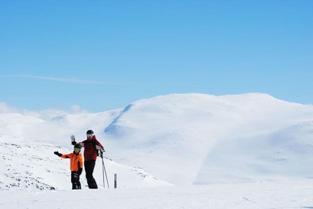 Due partecipanti a un viaggio di gruppo WeRoad, in tenuta da sci, in piedi su una montagna innevata, con uno che saluta, sotto un cielo azzurro e limpido.