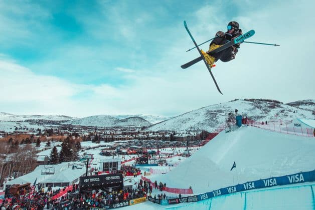 Un esquiador acrobatico realiza un truco aereo con los esquis cruzados sobre una rampa de nieve durante una competicion con una multitud abajo